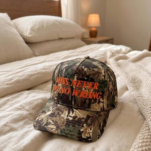 Peut inclure: Casquette de baseball camouflage avec le texte "MISS NEVER DO NO WRONG" en lettres orange. La casquette a une visière incurvée et une couronne structurée. Le chapeau est posé sur un lit avec une literie blanche.