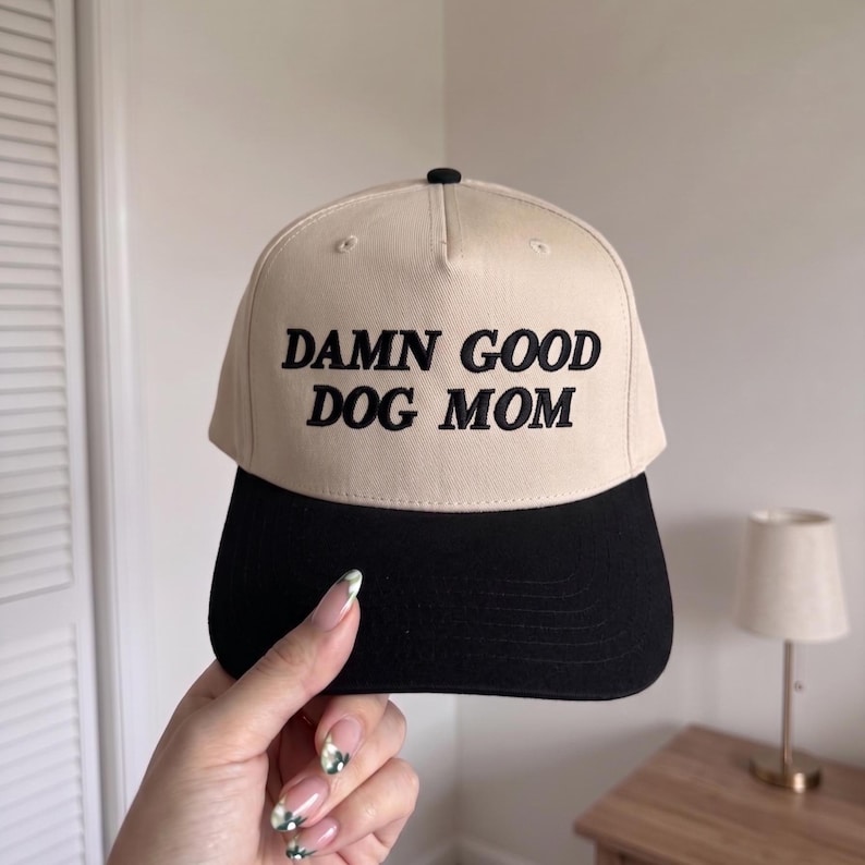 May include: A beige and black baseball cap featuring the embroidered text "DAMN GOOD DOG MOM". The cap has a curved peak and a button on top. A fashionable accessory for dog owners.