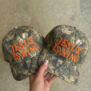 May include: Two camouflage baseball caps with the text "JESUS IS KING" embroidered in orange. The caps have a classic six-panel design and a curved brim. The camouflage pattern features shades of green, brown, and tan.