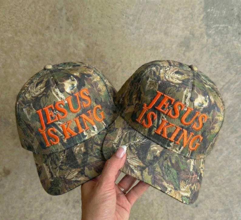 May include: Two camouflage baseball caps with the text "JESUS IS KING" embroidered in orange. The caps have a classic six-panel design and a curved brim. The camouflage pattern features shades of green, brown, and tan.