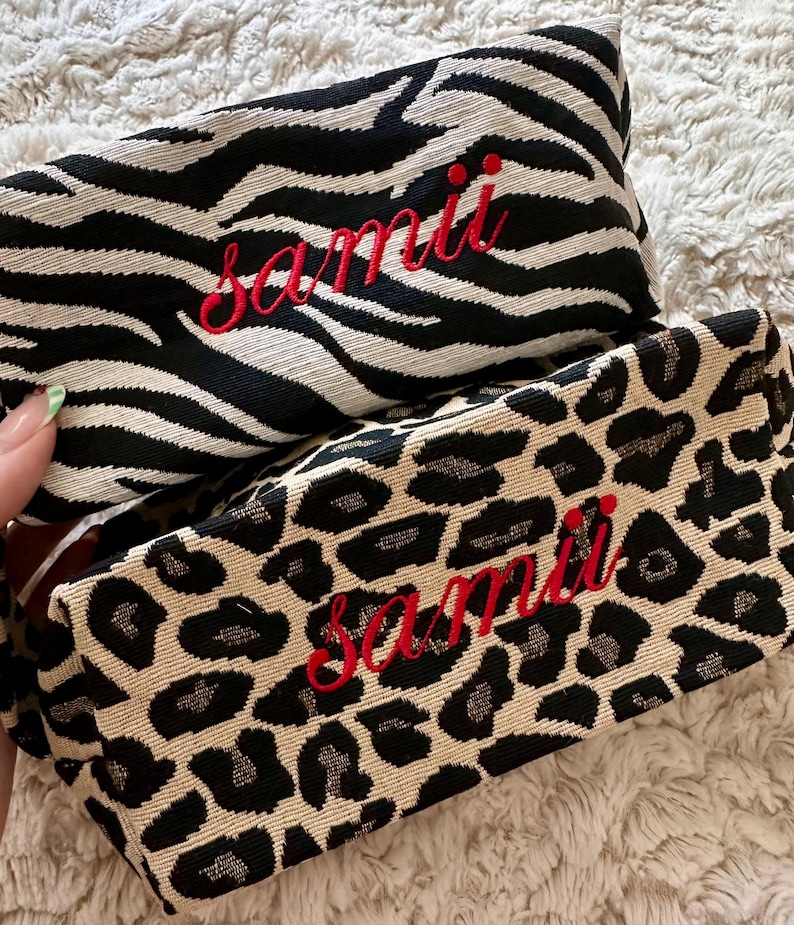 May include: Two rectangular cosmetic bags with animal print patterns. One features a black and white zebra stripe design, and the other has a tan and black leopard print. Both bags have the name "samii" embroidered in red.
