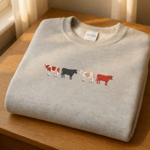 May include: A light gray sweatshirt with a crew neck. The sweatshirt features an embroidered design of four cows in a row, each in a different color: red and white, black, white and brown, and red.