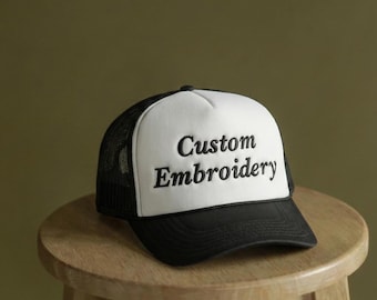 May include: A black and white trucker hat with the words "Custom Embroidery" embroidered on the front. The hat has a white front panel, a black brim, and a black mesh back. The hat is sitting on a wooden stool.