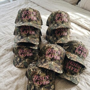 May include: A collection of camouflage baseball caps with the text "JESUS SAVES" in pink lettering. The caps are stacked and arranged on a bed, showcasing the hats' design and message.