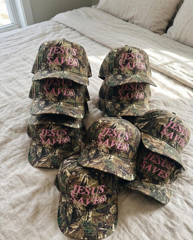 May include: A collection of camouflage baseball caps with the text "JESUS SAVES" in pink lettering. The caps are stacked and arranged on a bed, showcasing the hats' design and message.