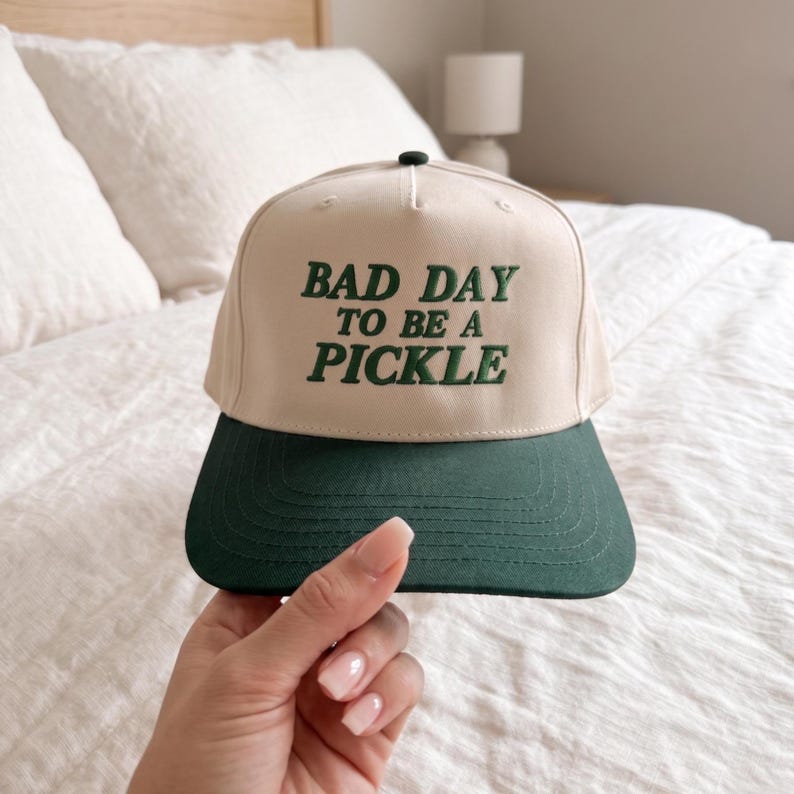 May include: A beige and dark green baseball cap featuring the text "BAD DAY TO BE A PICKLE" in green. The cap has a dark green peak and a black button. A casual hat.