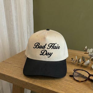 May include: A cream-colored baseball cap with a black brim. The cap features the embroidered phrase "Bad Hair Day" in black script. The hat is sitting on a wooden surface.