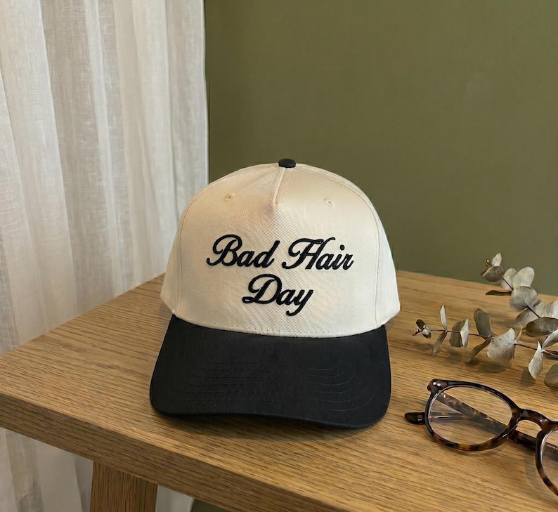 May include: A cream-colored baseball cap with a black brim. The cap features the embroidered phrase "Bad Hair Day" in black script. The hat is sitting on a wooden surface.