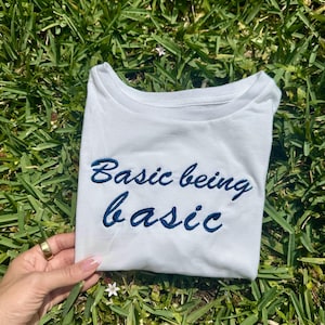 Op de afbeelding: Wit T-shirt met blauwe geborduurde tekst die "Basic being basic" luidt.