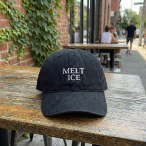 Può includere: Cappellino da baseball nero con la scritta "MELT ICE" ricamata in bianco. Il cappellino è realizzato in un tessuto morbido e strutturato e si trova su un tavolo di legno. Lo sfondo include un muro di mattoni e vegetazione.