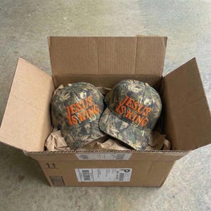 May include: Two camouflage baseball caps with the text "JESUS IS KING" embroidered in orange thread are inside a cardboard box. The caps have a curved brim and a six-panel design. The box is open, revealing the caps nestled in brown packing paper.