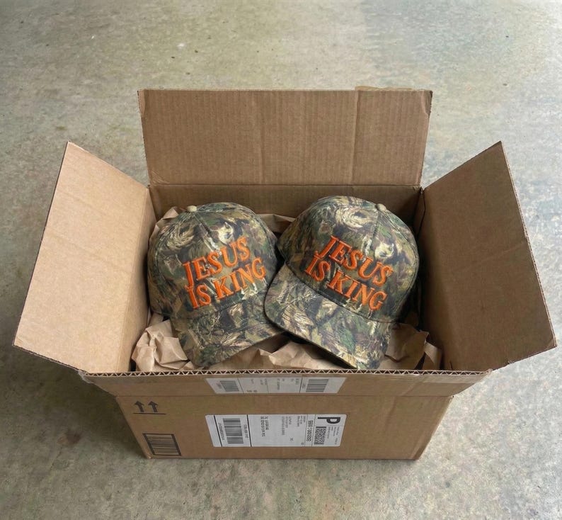 May include: Two camouflage baseball caps with the text "JESUS IS KING" embroidered in orange thread are inside a cardboard box. The caps have a curved brim and a six-panel design. The box is open, revealing the caps nestled in brown packing paper.