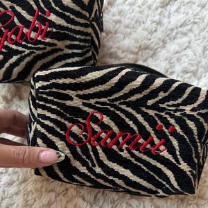Puede incluir: Dos estuches rectangulares con cremallera que presentan un patr&oacute;n de rayas de cebra en negro y beige. Cada estuche est&aacute; bordado con un nombre en letra cursiva roja, uno dice Gabi y el otro Samii, con un cierre de cremallera negro.