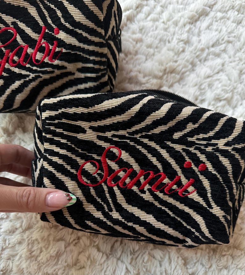 Puede incluir: Dos estuches rectangulares con cremallera que presentan un patr&oacute;n de rayas de cebra en negro y beige. Cada estuche est&aacute; bordado con un nombre en letra cursiva roja, uno dice Gabi y el otro Samii, con un cierre de cremallera negro.