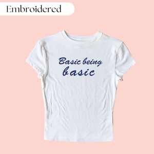 Könnte beinhalten: Weißes T-Shirt mit kurzen Ärmeln und Rundhalsausschnitt. Das Shirt hat einen blauen gestickten Text, der "Basic being basic" lautet.