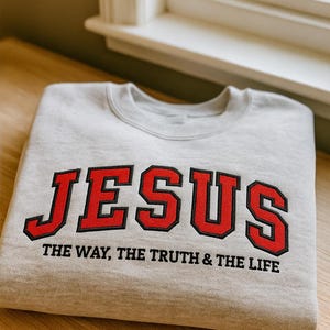 Könnte beinhalten: Ein hellgraues Sweatshirt mit dem Wort "JESUS" in großen, roten Buchstaben im College-Stil. Darunter steht der Satz "THE WAY, THE TRUTH & THE LIFE" in schwarzer Schrift. Das Sweatshirt liegt gefaltet auf einer Holzoberfläche.