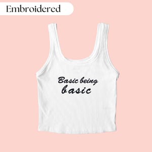 Op de afbeelding: Witte tanktop met de tekst "Basic being basic" geborduurd in zwart.