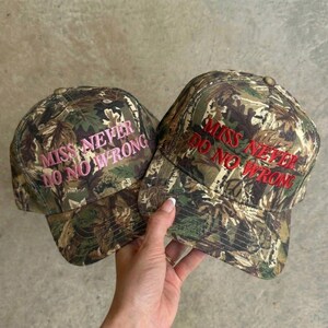 May include: Two camouflage baseball caps with the embroidered text "MISS NEVER DO NO WRONG". One cap has pink text, the other has red text. The caps are held by a person.