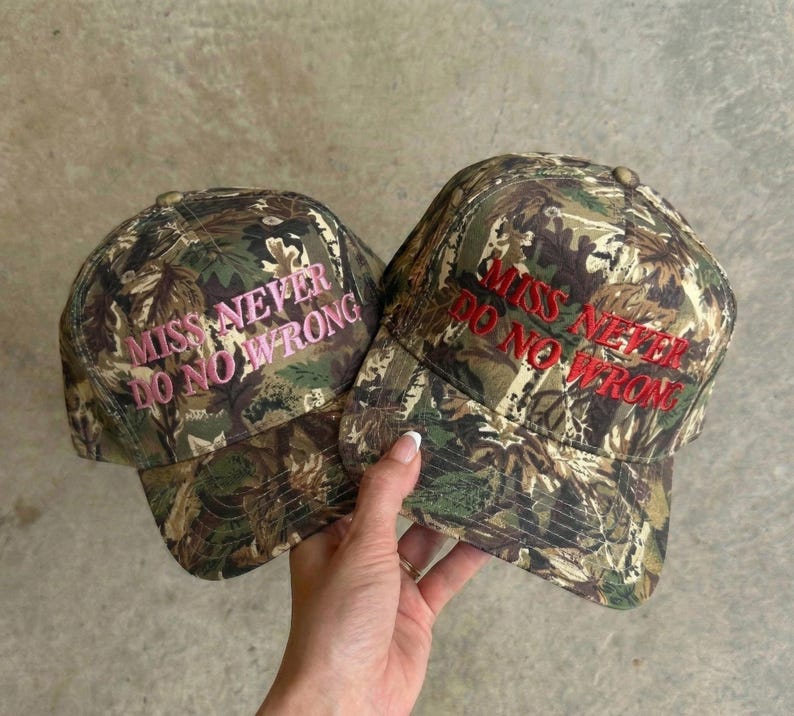 May include: Two camouflage baseball caps with the embroidered text "MISS NEVER DO NO WRONG". One cap has pink text, the other has red text. The caps are held by a person.