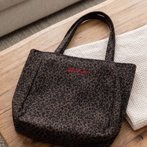 Op de afbeelding: Een donkerbruine en zwarte tote bag met luipaardprint en twee schouderbanden. De voorkant van de tas is voorzien van de geborduurde rode tekst "full of life" in het midden bij de bovenkant. De tas heeft een gestructureerde vorm.