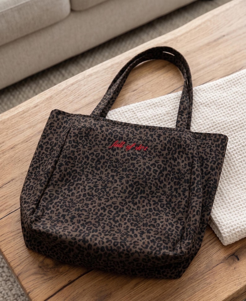 Op de afbeelding: Een donkerbruine en zwarte tote bag met luipaardprint en twee schouderbanden. De voorkant van de tas is voorzien van de geborduurde rode tekst "full of life" in het midden bij de bovenkant. De tas heeft een gestructureerde vorm.