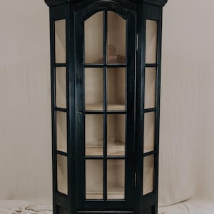 Puede incluir: Un mueble de esquina negro con puertas de cristal y un cajón. El mueble tiene una parte superior curvada y está pintado con un acabado mate oscuro. Las puertas de cristal revelan estantes interiores. Un pequeño pomo redondo de latón adorna el cajón.