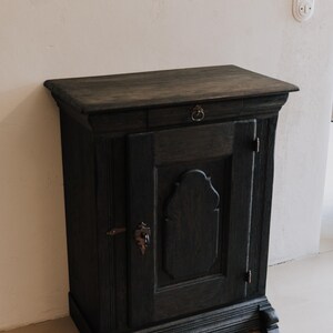 May include: Dark stained wooden cabinet with a single drawer and a door. The cabinet has ornate details, including a decorative escutcheon and a ring pull. The cabinet is set against a neutral wall.