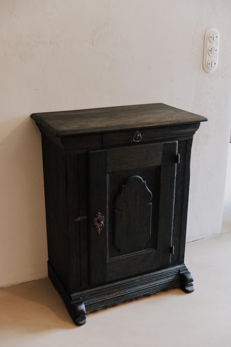 May include: Dark stained wooden cabinet with a single drawer and a door. The cabinet has ornate details, including a decorative escutcheon and a ring pull. The cabinet is set against a neutral wall.