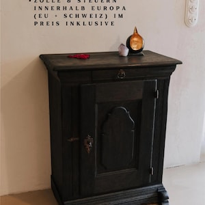 May include: A dark stained wooden cabinet with a single door and a small drawer. The cabinet has decorative molding and a metal handle. A small, gold-colored object sits on top, along with a crystal and a red object.