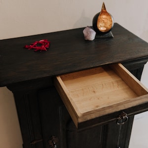 May include: Dark stained wooden cabinet with an open drawer. The cabinet has a rectangular top with a small arrangement of red petals, a purple crystal, and a decorative candle holder. The drawer's interior is light-colored wood.