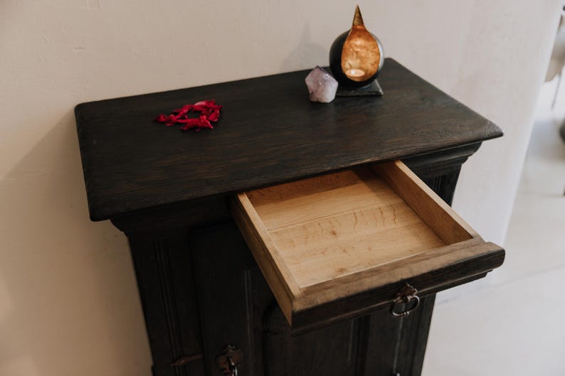 May include: Dark stained wooden cabinet with an open drawer. The cabinet has a rectangular top with a small arrangement of red petals, a purple crystal, and a decorative candle holder. The drawer's interior is light-colored wood.