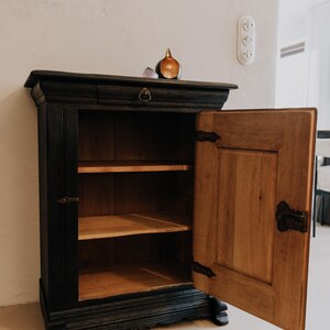 May include: A black wooden cabinet with an open door revealing two shelves. The cabinet has a drawer with a round handle and decorative hardware. A small decorative object sits on top.