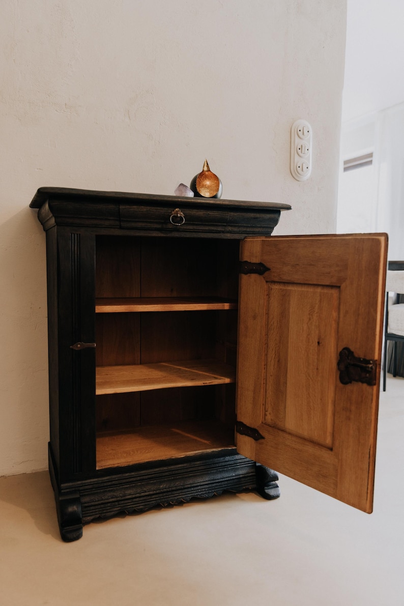 May include: A black wooden cabinet with an open door revealing two shelves. The cabinet has a drawer with a round handle and decorative hardware. A small decorative object sits on top.