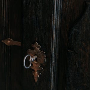 May include: Close-up of a dark wooden door with a vintage, ornate keyhole and key. The keyhole and key are made of aged, rusty metal. The door's wood grain is visible, with vertical lines and a dark brown color.