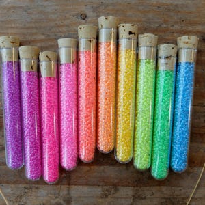 May include: Ten glass test tubes filled with colorful beads. The tubes have cork tops and are arranged in a rainbow pattern from purple to blue.