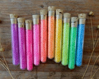 May include: Ten glass test tubes filled with colorful beads. The tubes have cork tops and are arranged in a rainbow pattern from purple to blue.
