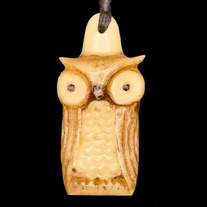 Puede incluir: Un colgante de madera tallada en forma de búho, de color marrón claro y crema. El búho tiene ojos grandes y redondos y un pico detallado.