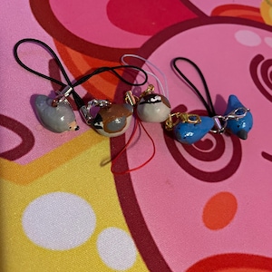 Puede incluir: Cuatro pequeños amuletos con forma de pájaro en varios colores. Los amuletos están unidos a lazos negros o blancos. Los pájaros están pintados en tonos grises, marrones y azules, con detalles como picos y ojos. El fondo es una tela colorida estampada.