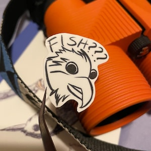 May include: A white sticker with a cartoon bird head and the text "FISH??" is held by tweezers. The sticker is in front of orange binoculars with a black strap. The background is a blurred image of a drawing.