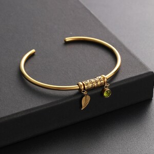 May include: Gold bangle bracelet with a charm that has the names "Alvic", "Nath", "Vden", "Julie" engraved on it. The charm has a small green gemstone and a leaf charm hanging from it.