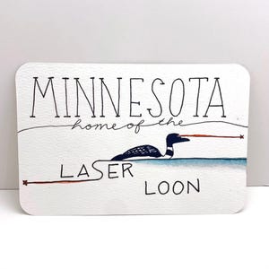 May include: A white card with black handwritten text that reads "Minnesota, home of the laser loon". A cartoon loon with a red laser beam coming from its eyes is illustrated on the card.