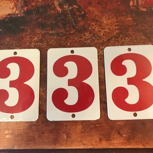 May include: Three rectangular white metal signs with the number 3 in red. Each sign has a hole at the top for hanging. The signs are arranged in a row.