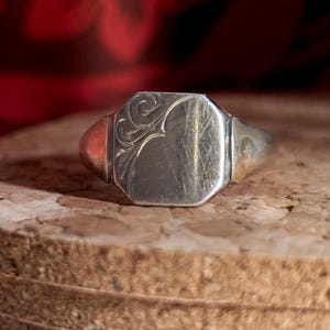 May include: A silver signet ring with an octagonal face and decorative flourishes. The ring has a rectangular face with a detailed floral design on one side. The band and shoulders of the ring are a contrasting gold color.