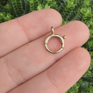 May include: A small, circular gold-toned spring ring clasp featuring a smooth, polished metal surface. The jewelry component includes a tiny attached loop at the top and a small protruding lever mechanism on the side for opening and closing.
