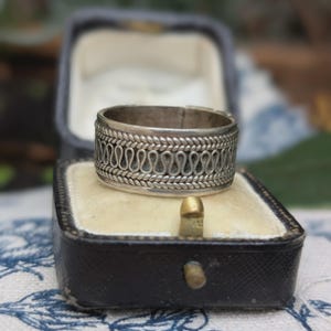 May include: A silver ring with a detailed pattern of loops and braided edges, displayed in a vintage black jewelry box with a cream-colored interior. The ring's design is intricate and eye-catching.