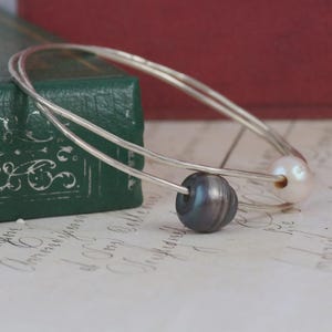 May include: A silver bangle bracelet featuring two distinct beads threaded onto the wire. One bead is a dark, iridescent charcoal grey, while the other is a pale, soft pink. The metal band has a smooth, polished finish and a circular shape.