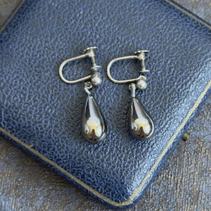 May include: A pair of silver-toned screw-back earrings featuring smooth, polished, dark grey teardrop-shaped pendants. The earrings have a metallic finish with visible reflections and a simple, functional screw mechanism for securing the jewellery to the ear.