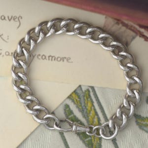 May include: A silver curb chain bracelet featuring thick, interlocking oval links with a polished metallic finish. The bracelet includes a secure lobster claw clasp mechanism and displays small, stamped hallmarks on each individual metal link.