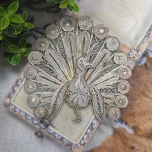 Pode incluir: Broche de pavão em filigrana prateada com cauda aberta, detalhes intrincados e cabeça de pássaro. O broche é exibido numa caixa de joias vintage com interior creme.