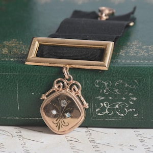 May include: A gold-toned brooch featuring a rectangular bar attached to a black fabric ribbon. A pendant hangs from the bar, displaying an engraved floral motif accented with three small, round, light-colored stones set in a central cluster.